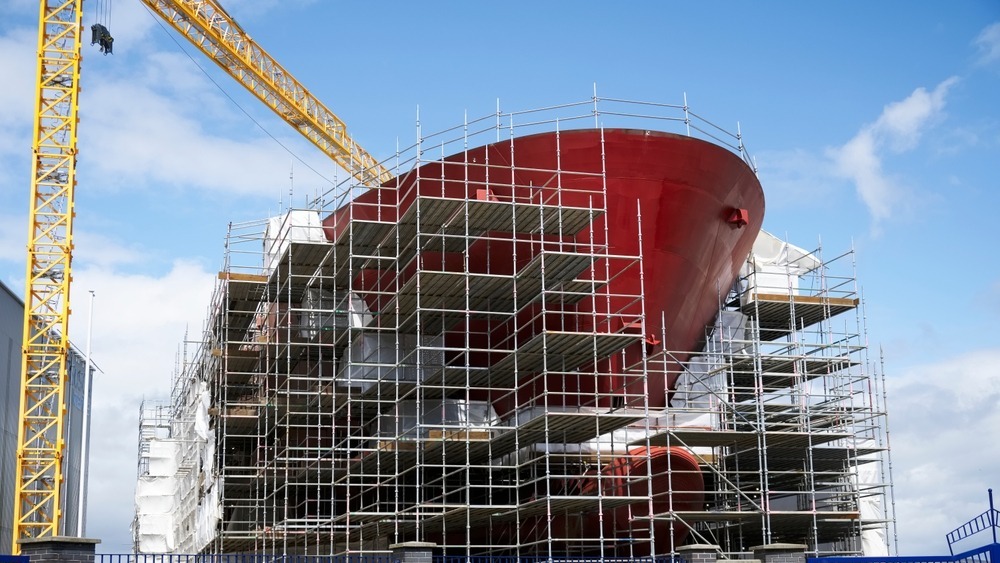 Skip med rødt skrog under bygging med stor kran til venstre.